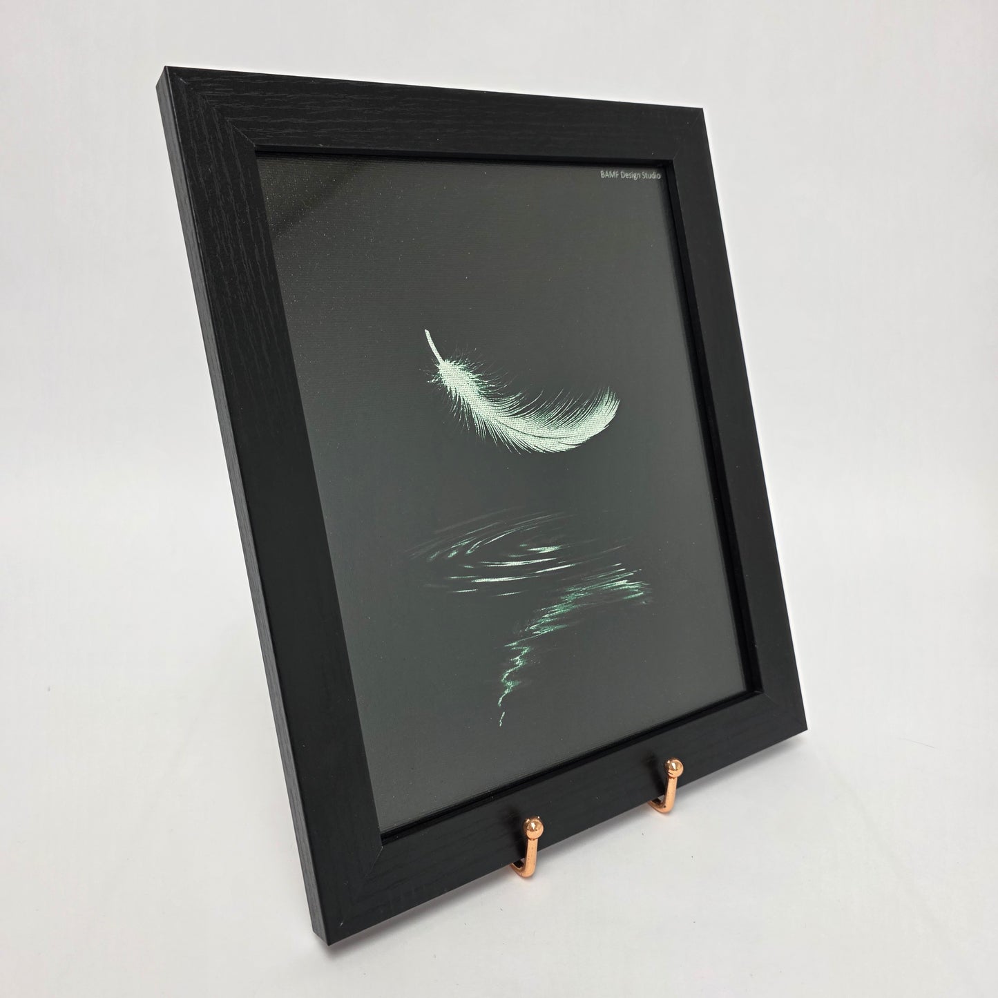 A framed canvas. Image of a green feather floating down with a ripple of water beneath it. Black background with green accents.