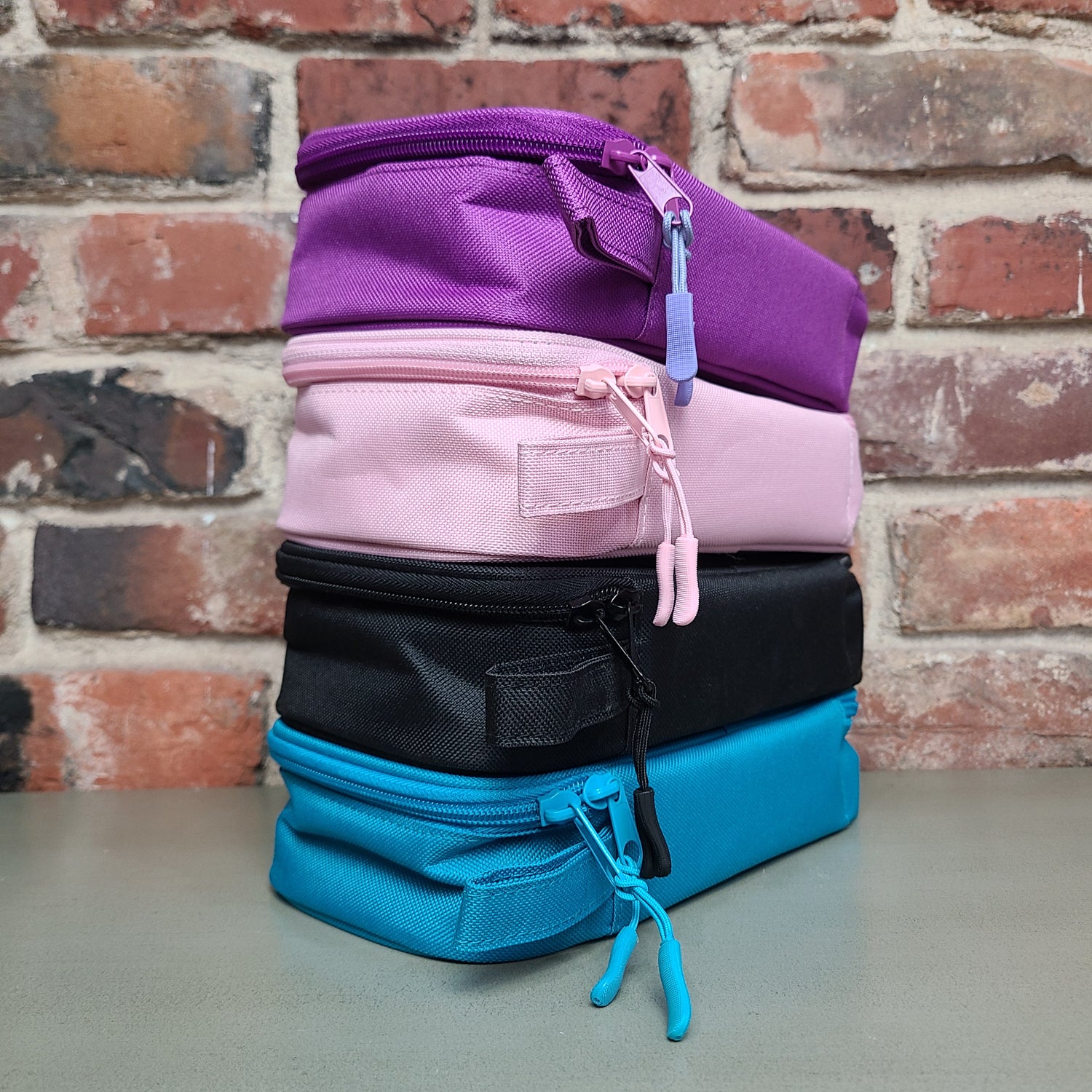 Image of four bags stacked on a surface in front of a brick wall. Bag colors include pink, black, blue, and purple.