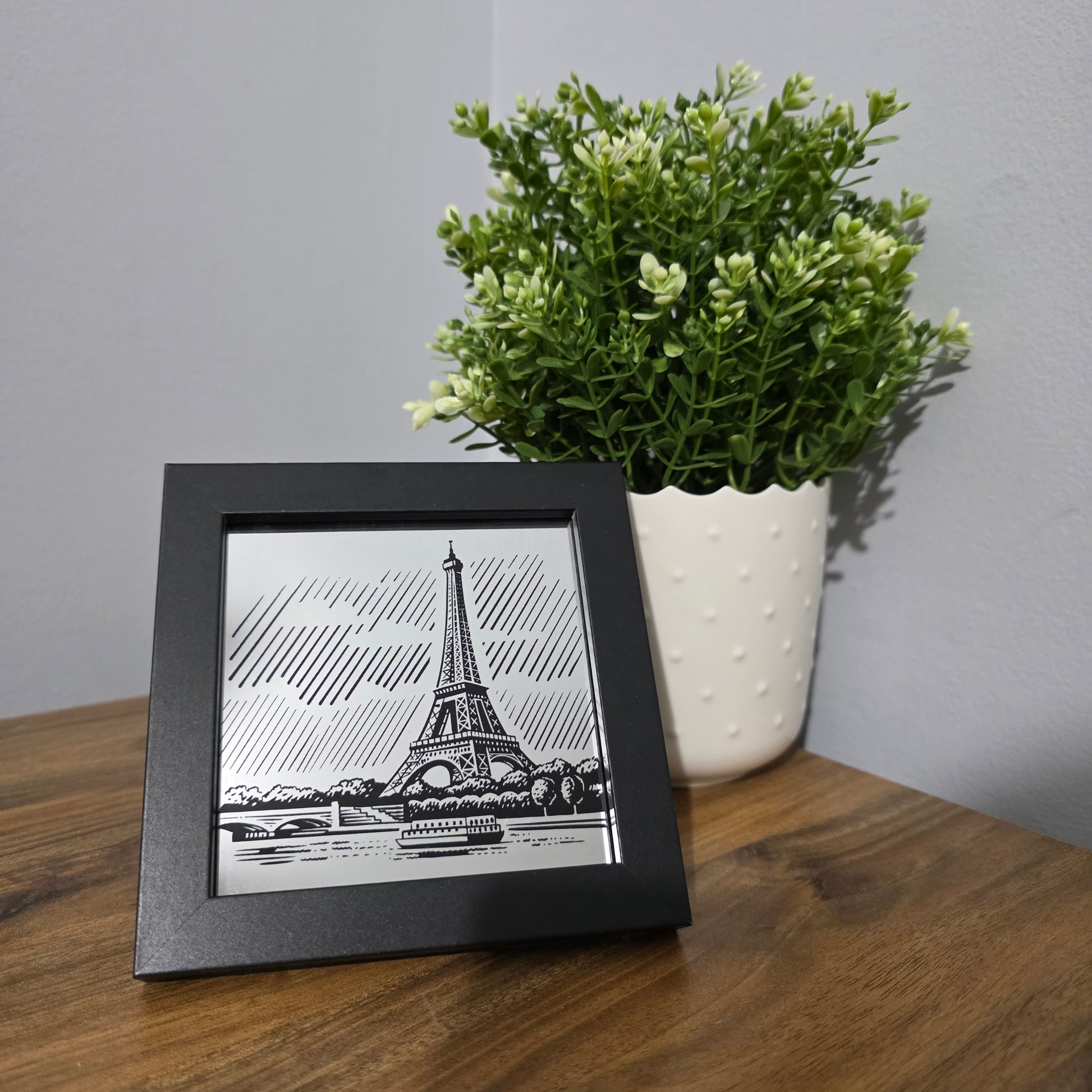 Framed glass artwork of the Eiffel Tower on a wooden surface with a plant.