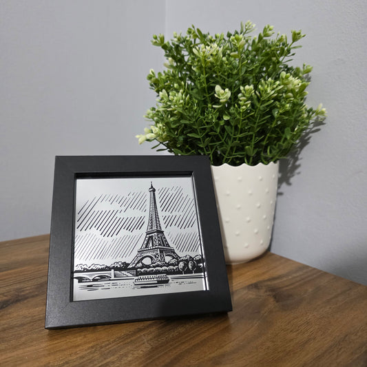 Framed glass artwork of the Eiffel Tower on a wooden surface with a plant.