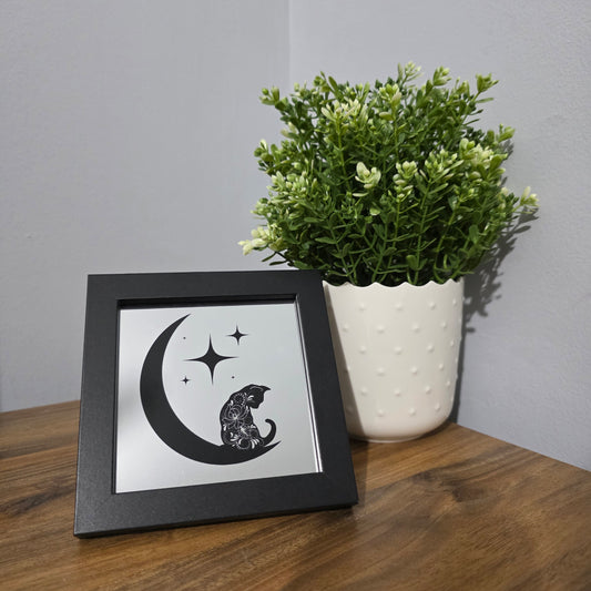 Framed glass artwork with a black design, sitting on a wooden surface next to a plant.