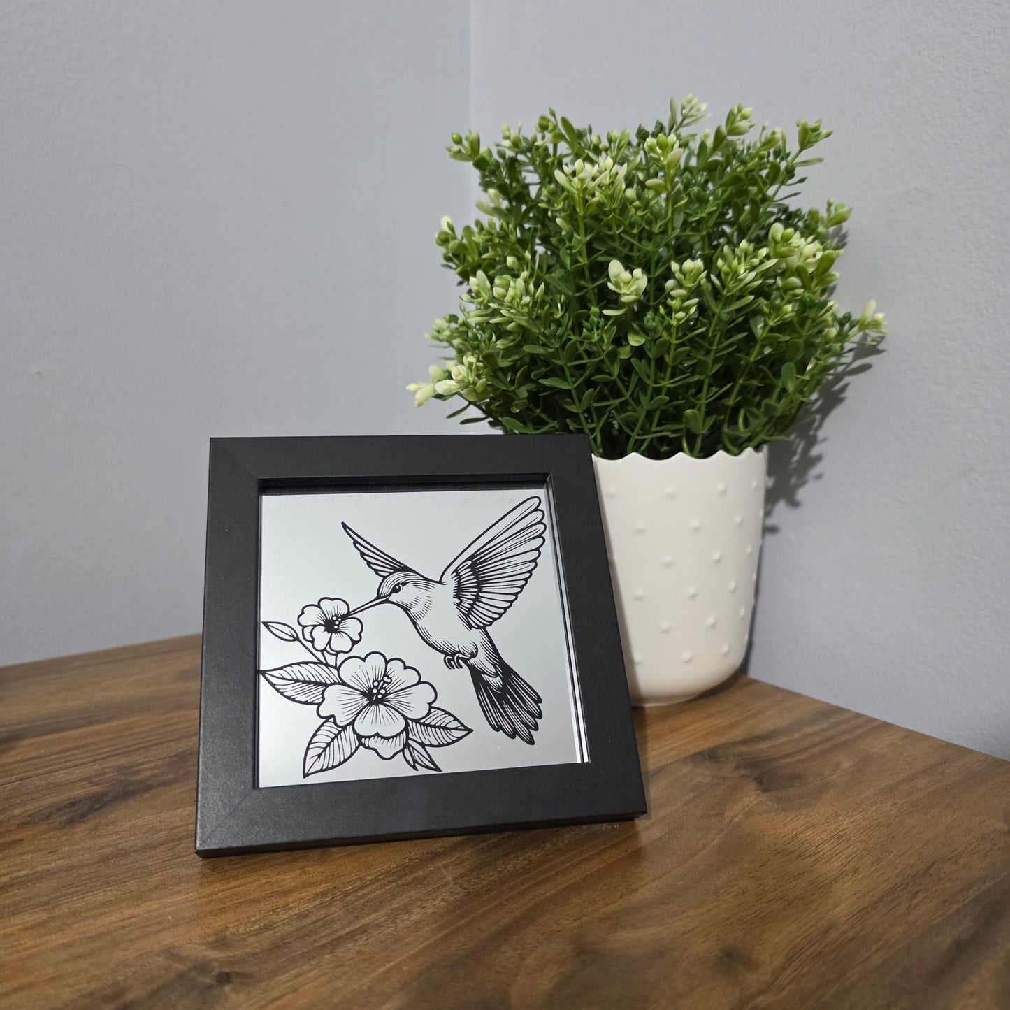 Framed black artwork of a bird and flower on a mirror, on a wooden surface with a plant.