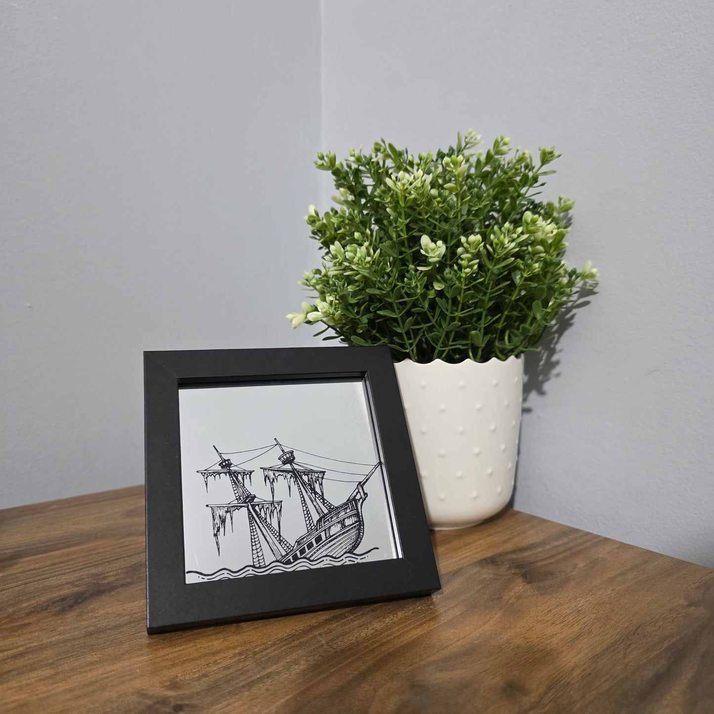 Framed glass art with black artwork of a sinking ship, sitting on a wooden surface next to a plant.