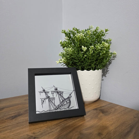 Framed glass art with black artwork of a sinking ship, sitting on a wooden surface next to a plant.